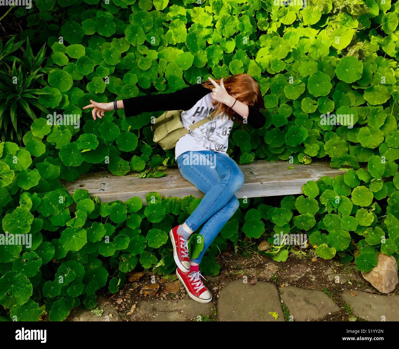 Preteen on a bench wearing red shoes in front of a wall of nasturtiums, hiding her face. Stock Photo