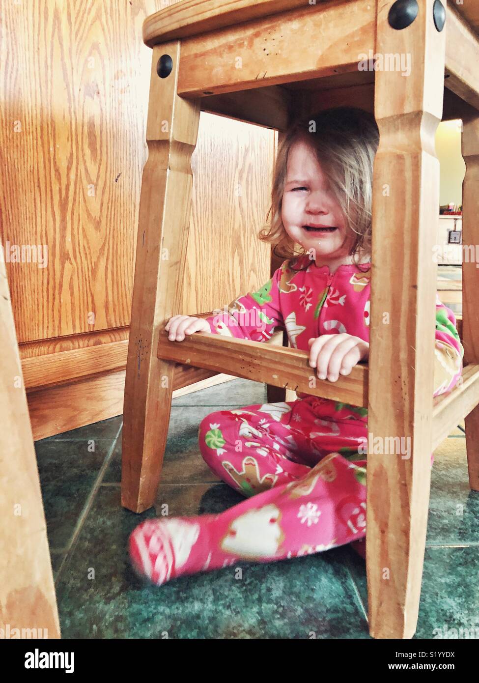Toddler girl in Christmas onesie pyjamas crying on tile floor under a rustic wooden stool Stock Photo
