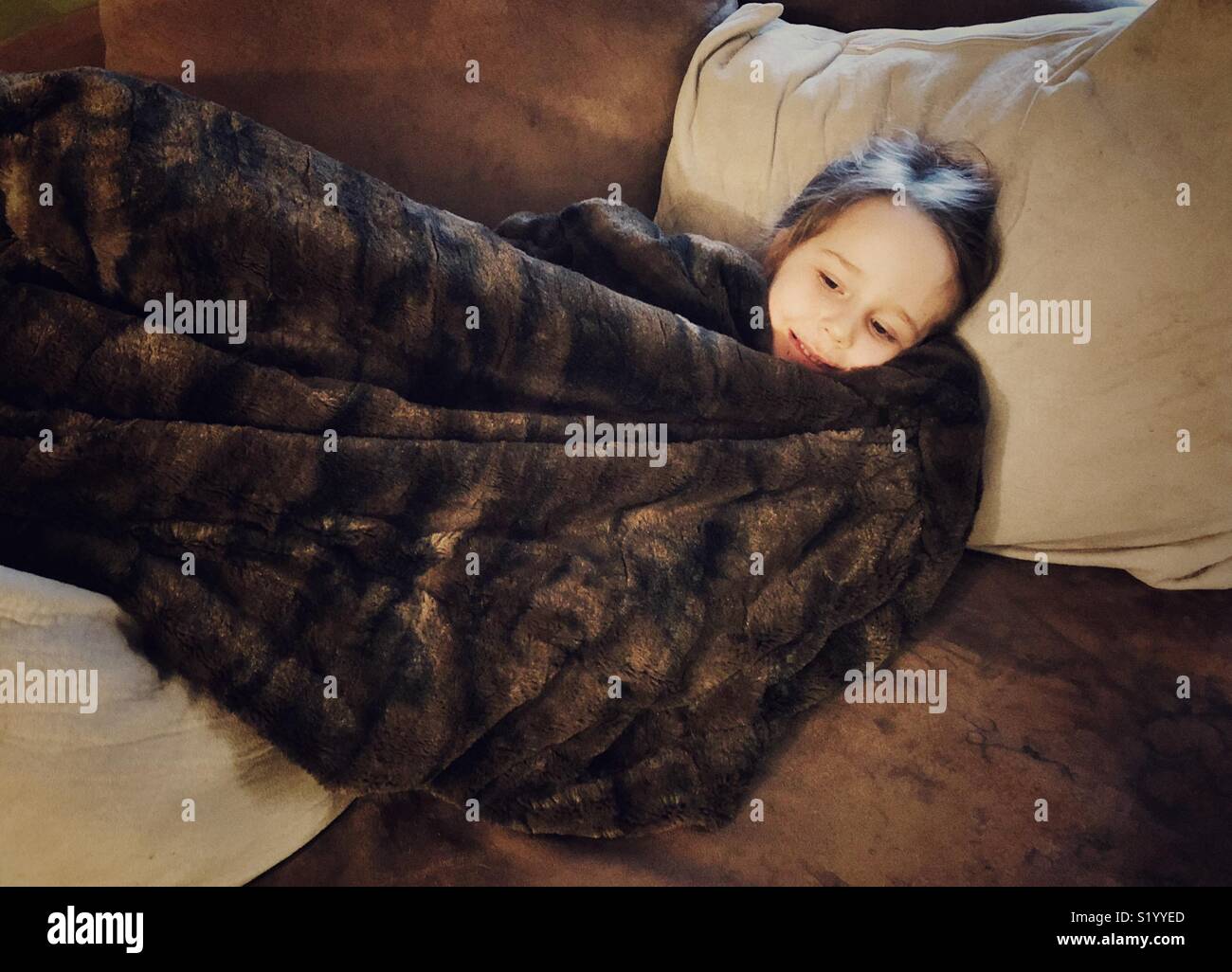 Young school age girl wrapped in brown faux fur blanket on cosy brown and beige sofa Stock Photo