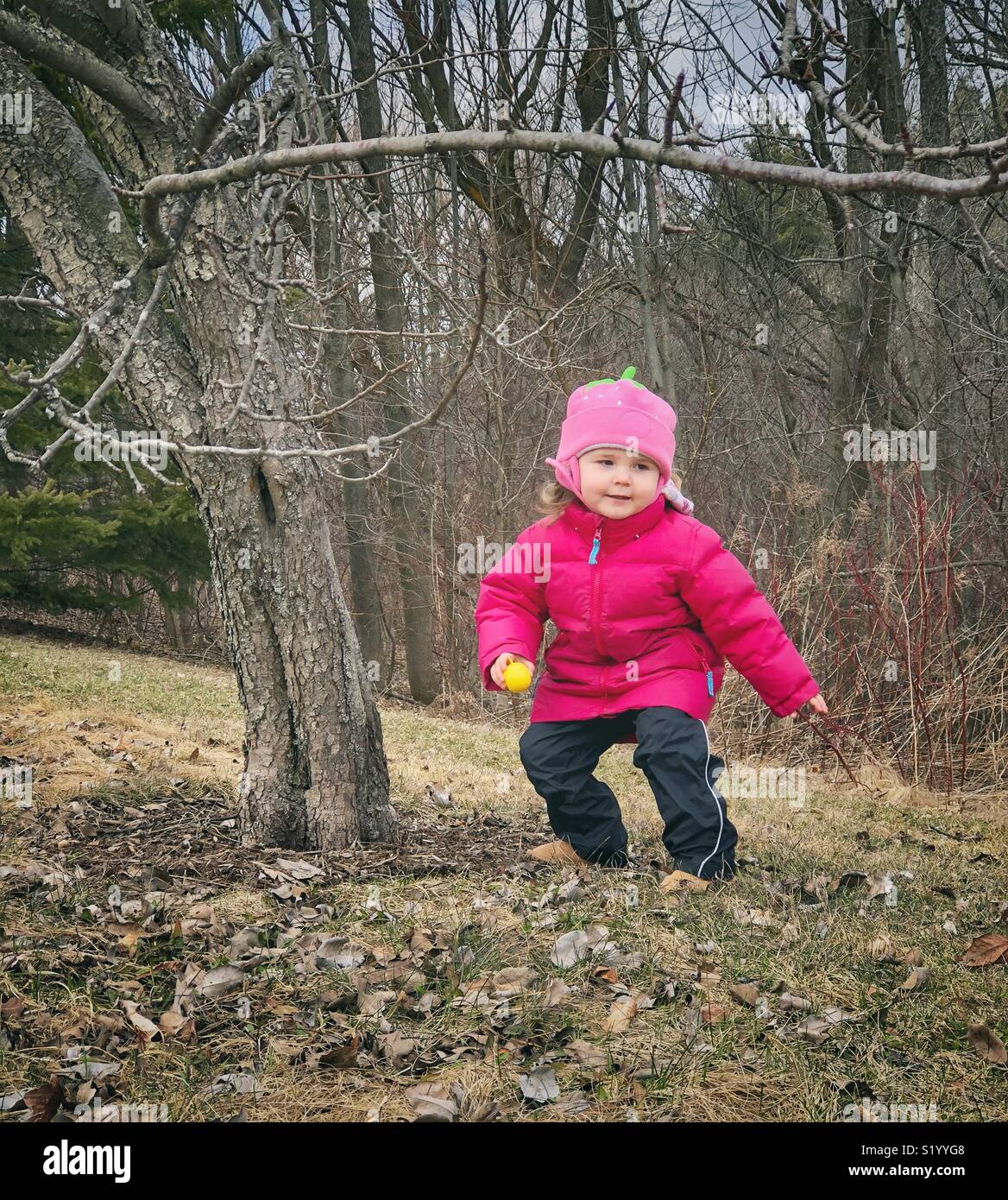 Toddler girl with plastic yellow Easter egg during hunt outdoors in cold weather - Smartphone Captured Stock Image
