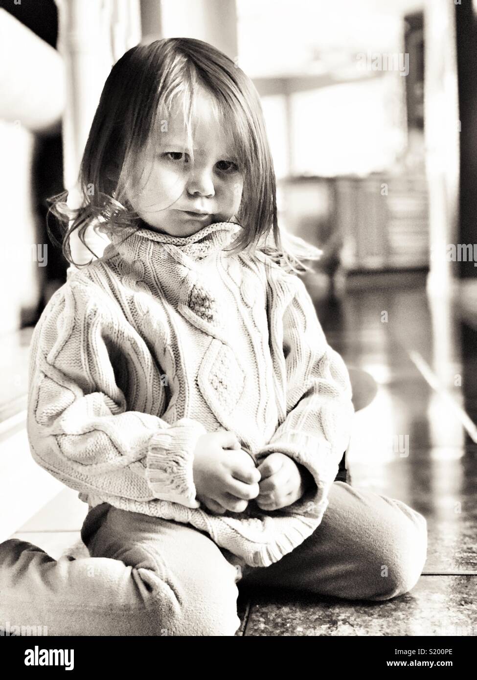 Black and white image of toddler girl looking sad and angry on tile floor with bright backlighting Stock Photo