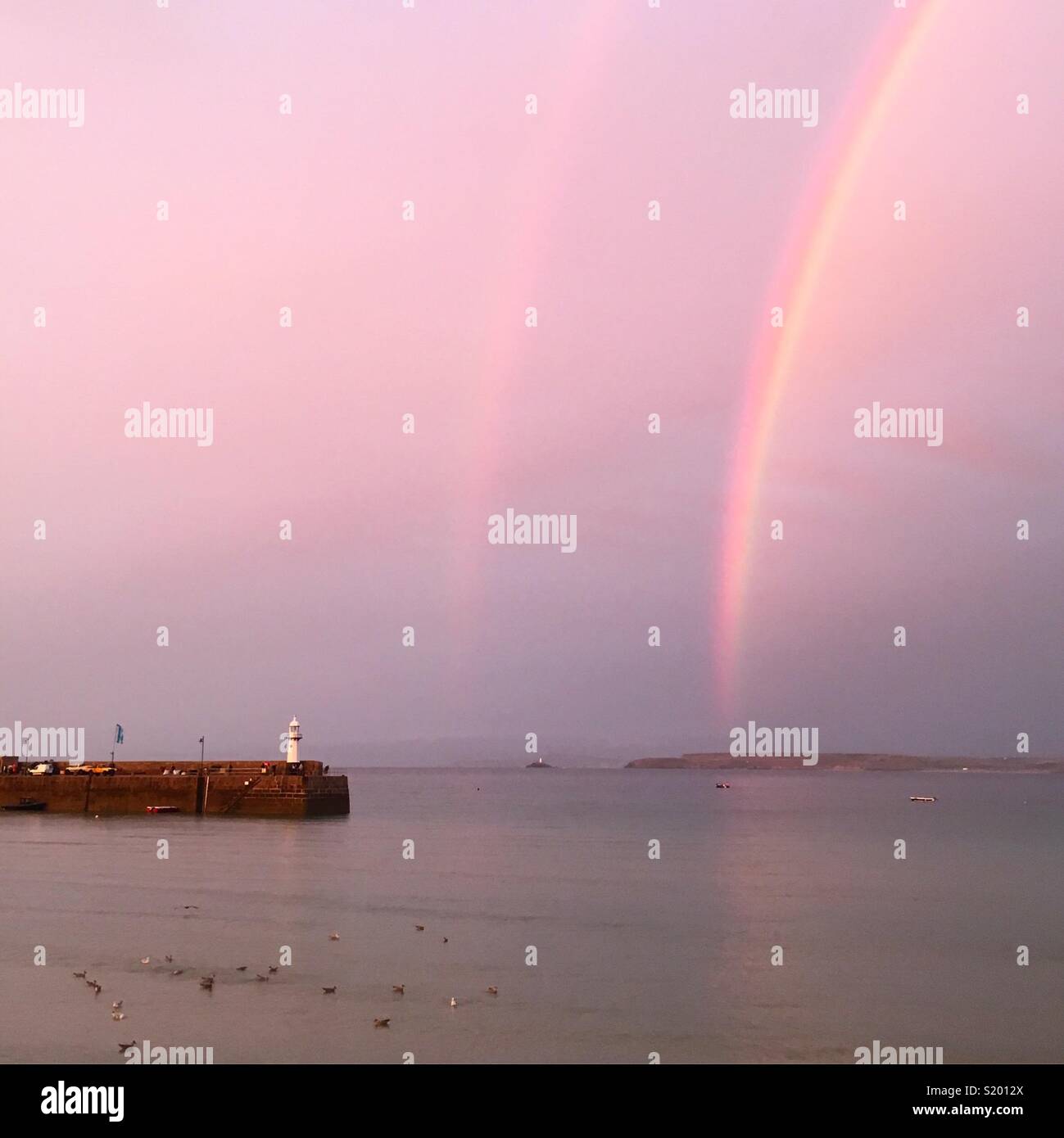 Double rainbow and pink sky in St Ives, Cornwall, looking out to sea, past the harbour wall and lighthouse Stock Photo