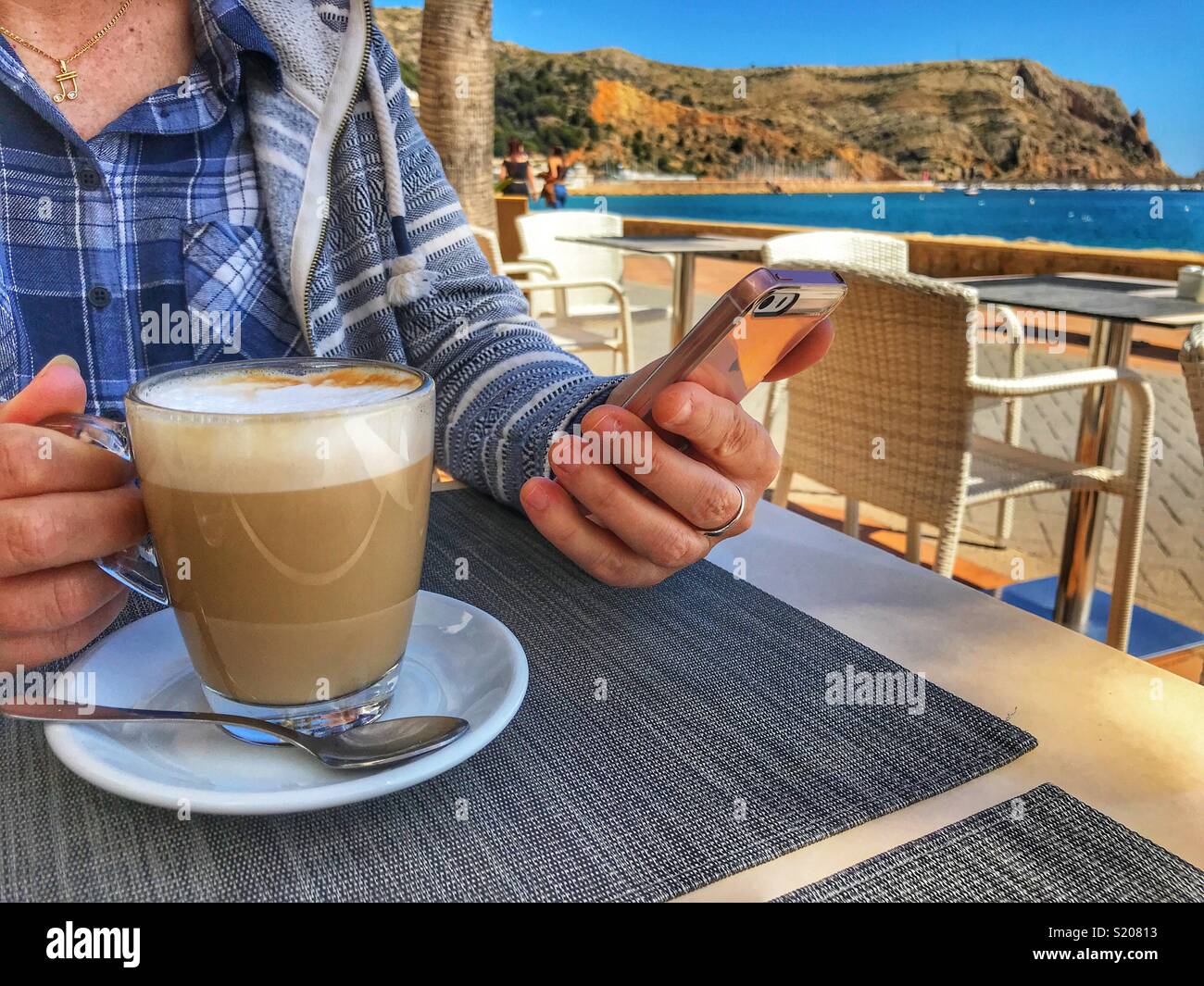 woman checking her mobile, with a cafe con leche, milky white coffee, on the waterfront promenade in the port area of Javea / Xabia on the Costa Blanca, Alicante Province, Comunidad Valenciana, Spain - Smartphone Captured Stock Image woman checking her mobile, with a cafe con leche, milky white coffee, on the waterfront promenade in the port area of Javea / Xabia on the Costa Blanca, Alicante Province, Comunidad Valenciana, Spain - Smartphone Captured Stock Image