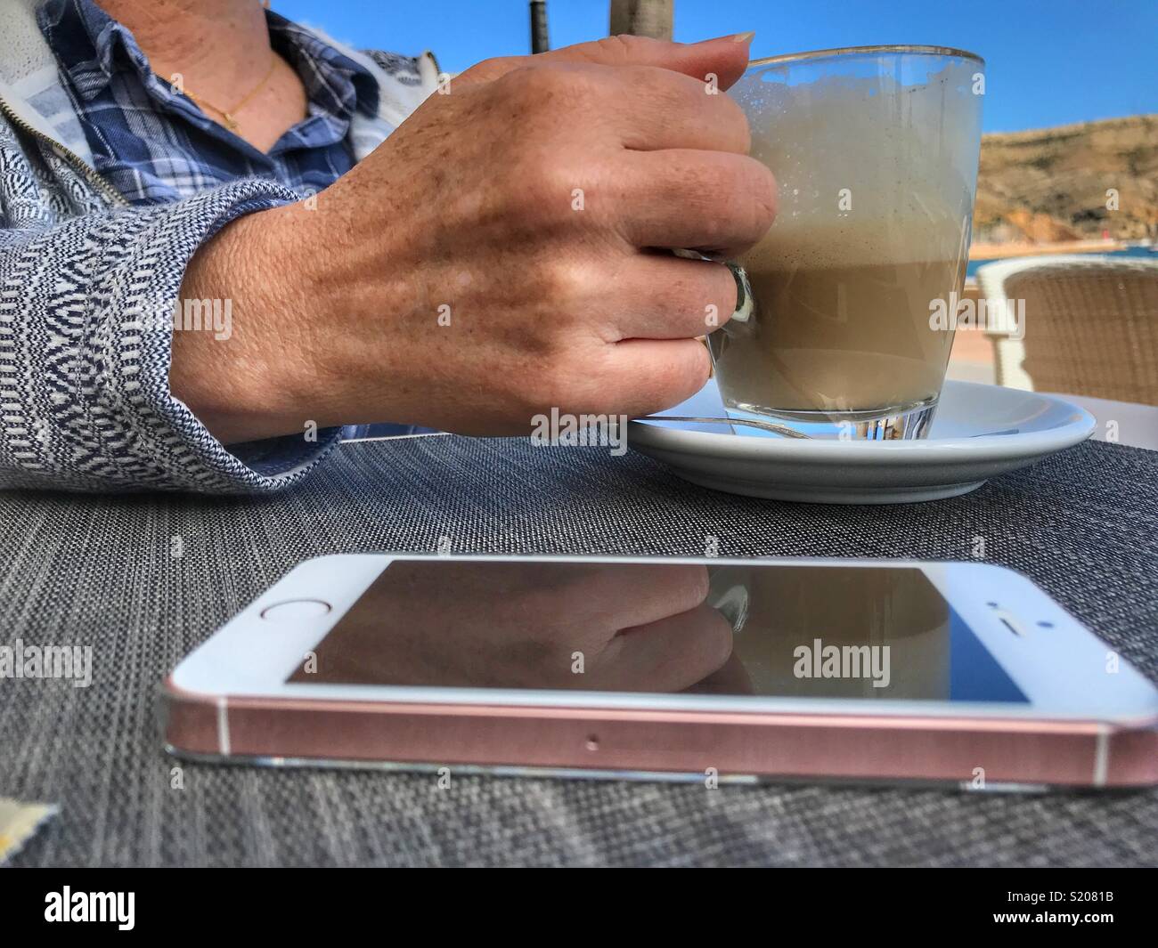 woman with a cafe con leche, milky white coffee, and smartphone on table, at waterfront cafe in the port area of Javea / Xabia on the Costa Blanca, Alicante Province, Comunidad Valenciana, Spain - Smartphone Captured Stock Image