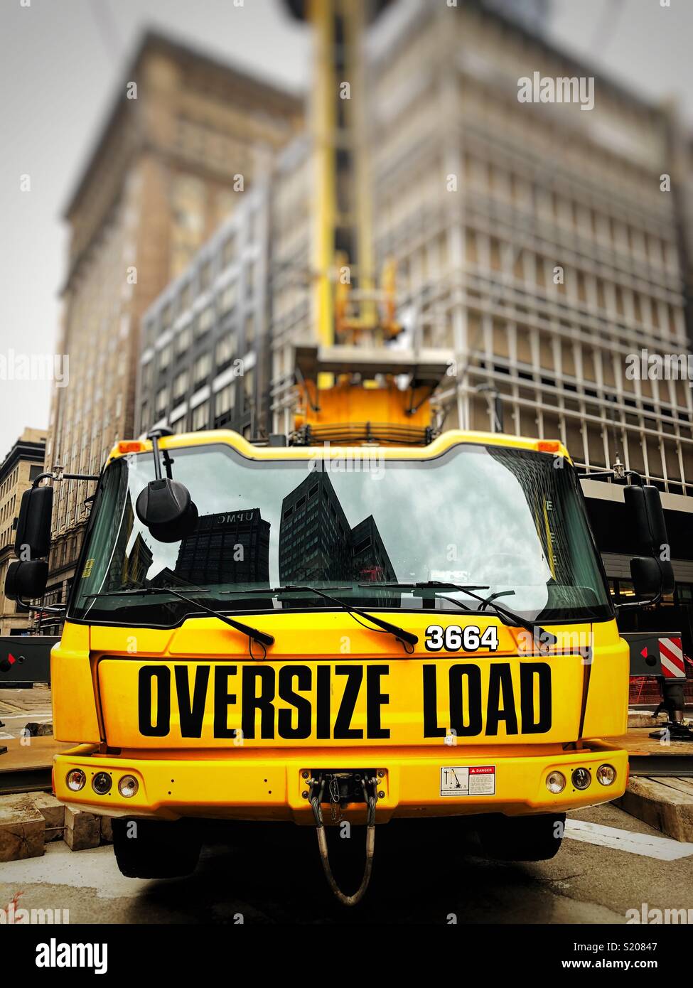 A large yellow crane labeled “oversize load” is parked on a city street corner in front of buildings - Smartphone Captured Stock Image