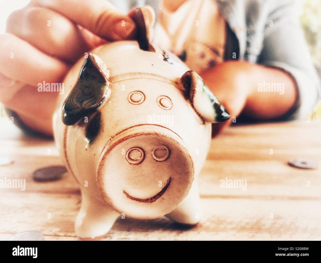 Millennial woman sitting at a table putting a coin into a piggy bank, midsection view - Smartphone Captured Stock Image