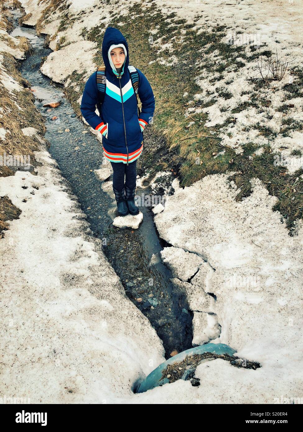 Girl standing on icy snow chunk in a ditch with flowing water Stock Photo