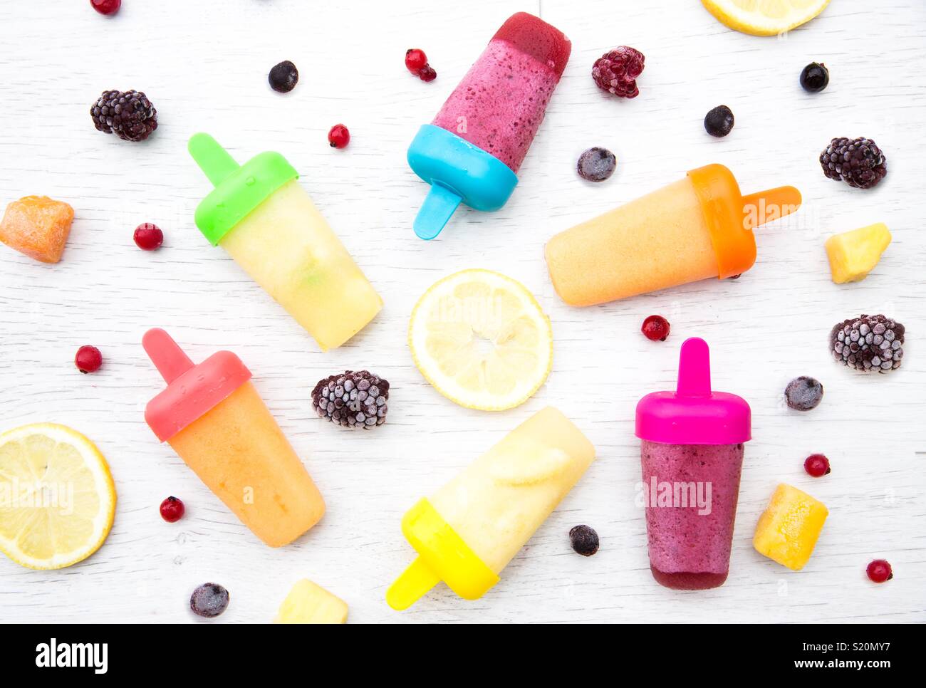 A flat lay food image of colourful fruit flavoured ice lollipops surrounded by fresh fruit ingredients on a white background. - Smartphone Captured Stock Image