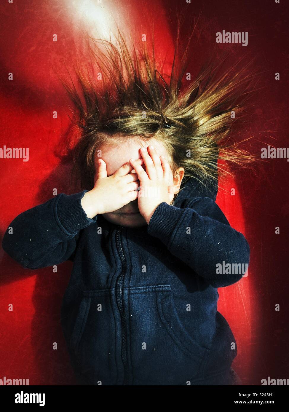 Toddler girl covering face with hands and hair sticking up due to static electricity from a red slide - Smartphone Captured Stock Image