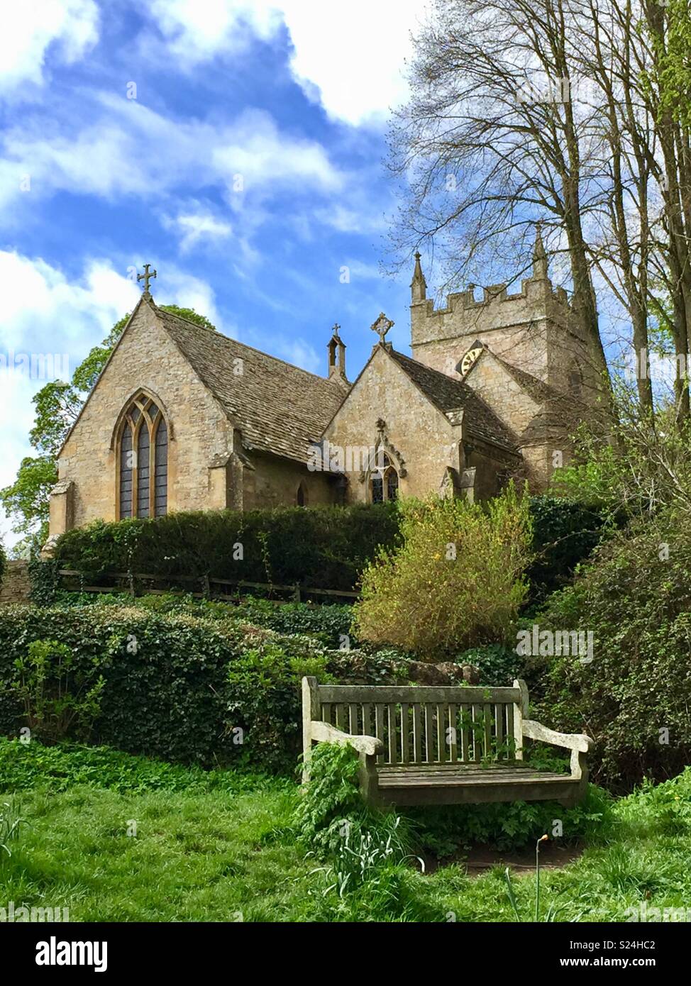 Church of St Peters in the village of Upper Slaughter, Gloucestershire, United Kingdom, looking uphill with wooden bench in foreground against a blue sky Stock Photo