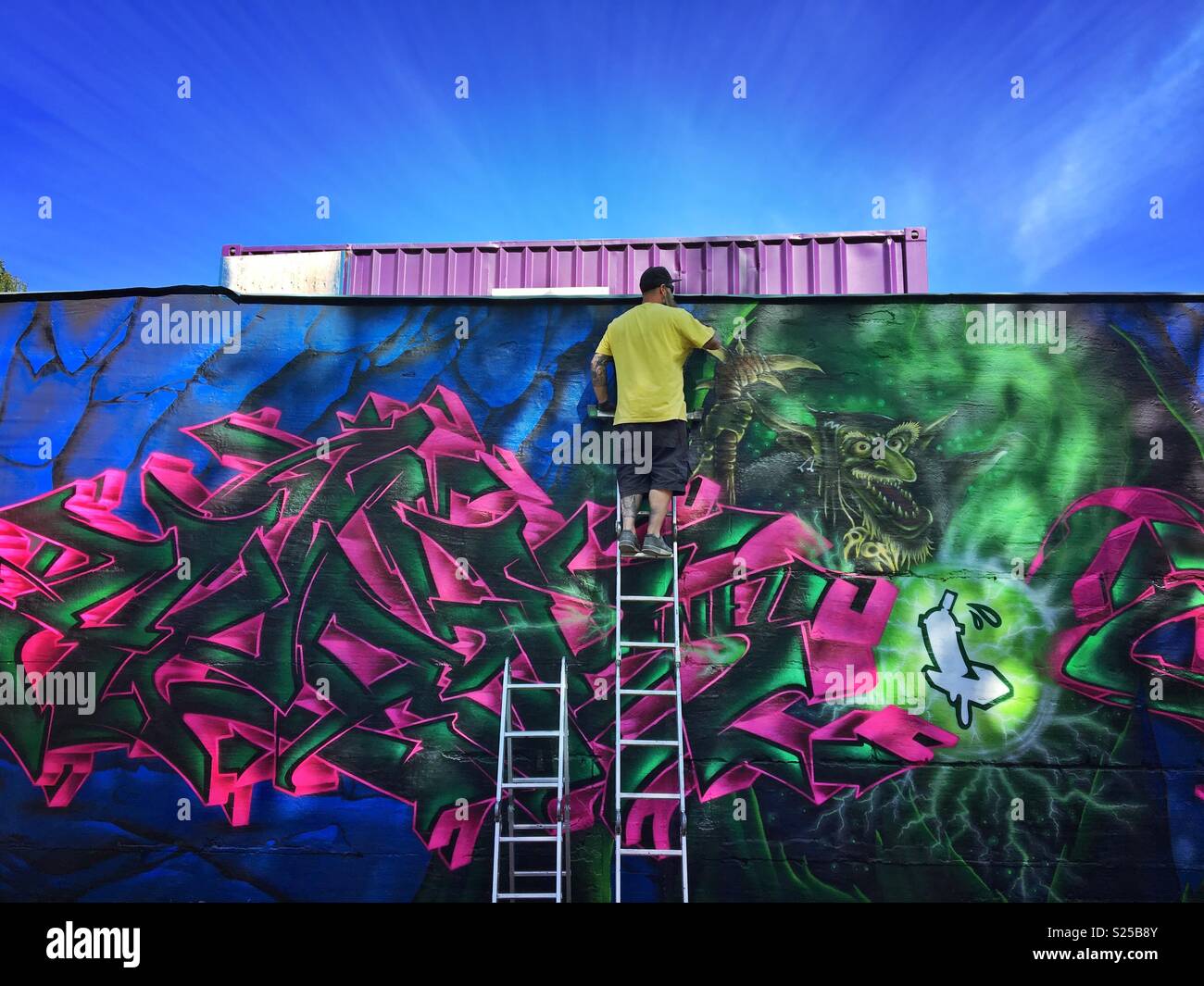Man spraying graffiti on wall in sunshine with ladder and blue sky - Smartphone Captured Stock Image