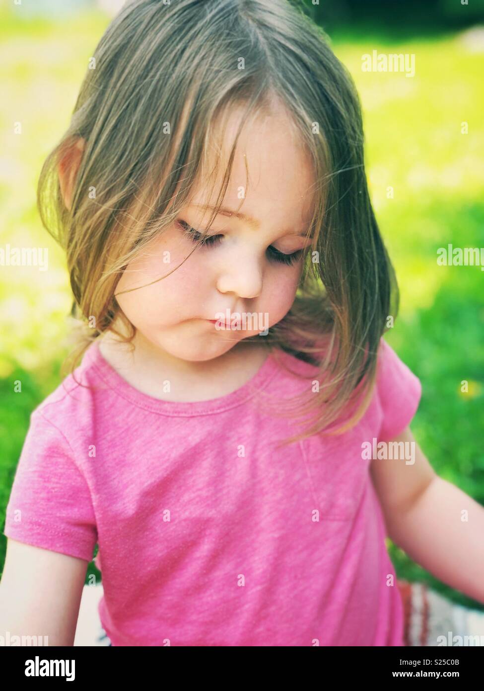 Toddler girl in pink t-shirt sitting outside looking down Stock Photo