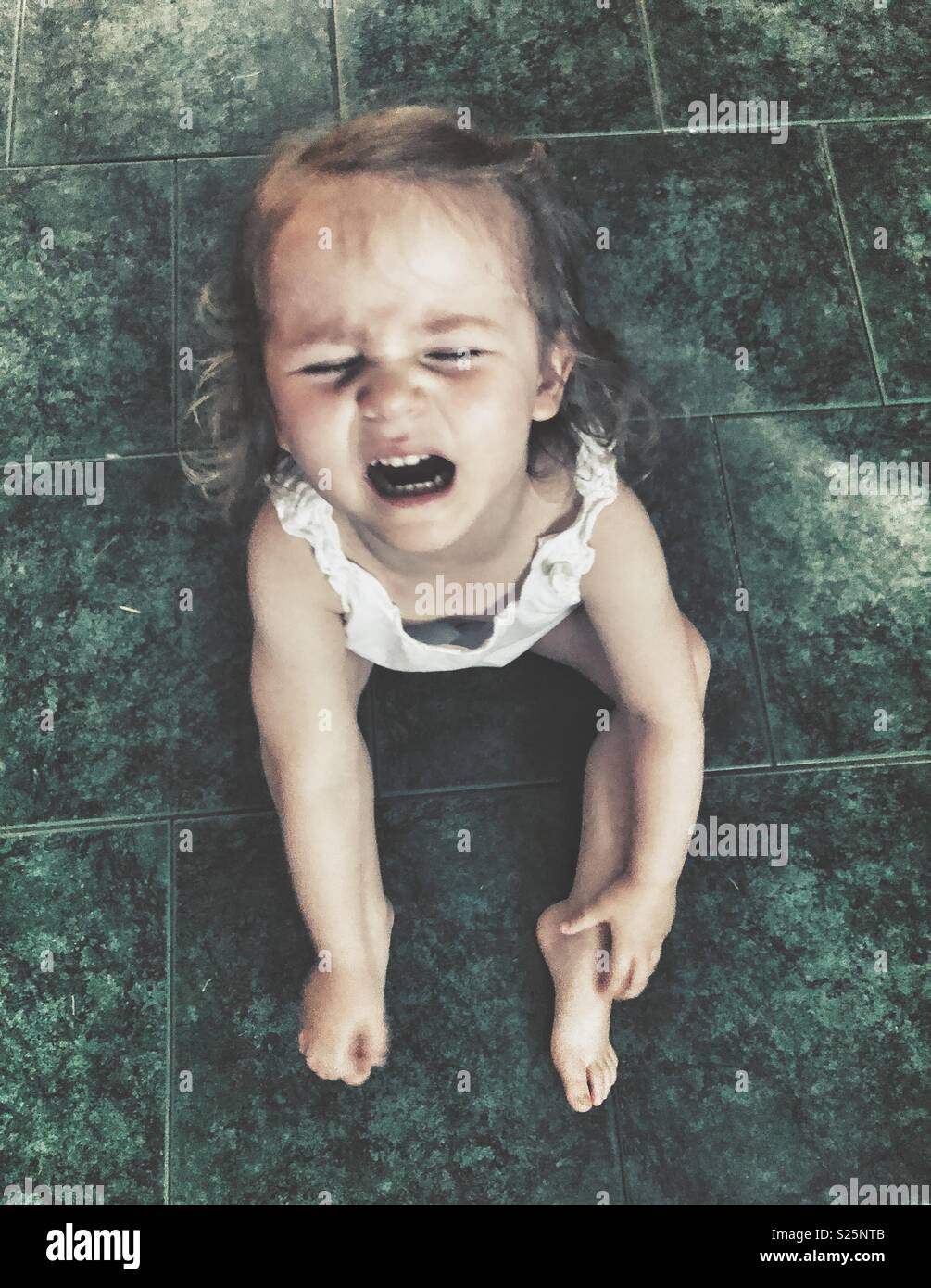 Toddler girl sitting on green tile floor crying during a temper tantrum Stock Photo