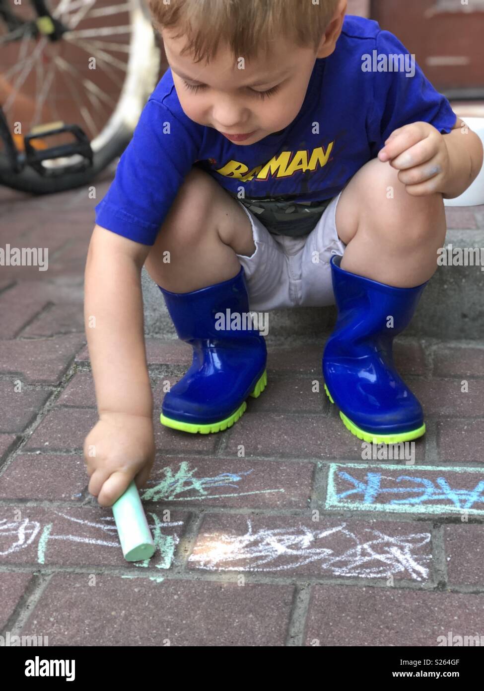 A boy coloring on a brick pavement outside with his rain shoes on Stock Photo