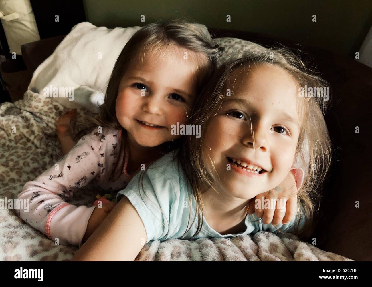 Sisters cuddling on a chair with a blanket happily smiling Stock Photo