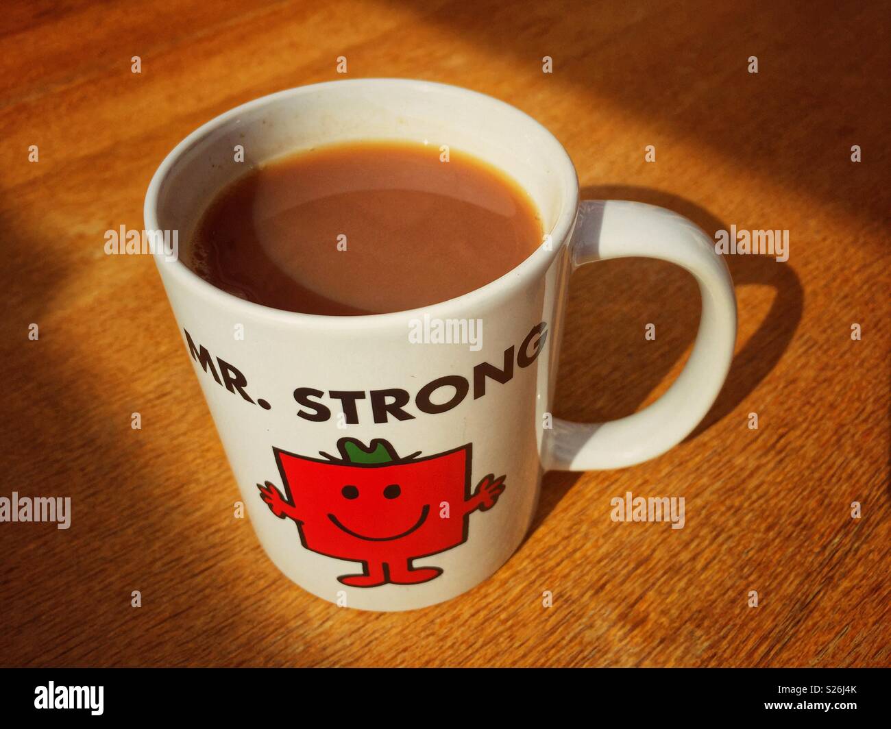 A white mug featuring Mr.Strong from the Mr. Men series, sitting on a wooden table bathed in sunlight, with a cozy, cheerful vibe. A strong cup of tea - Smartphone Captured Stock Image