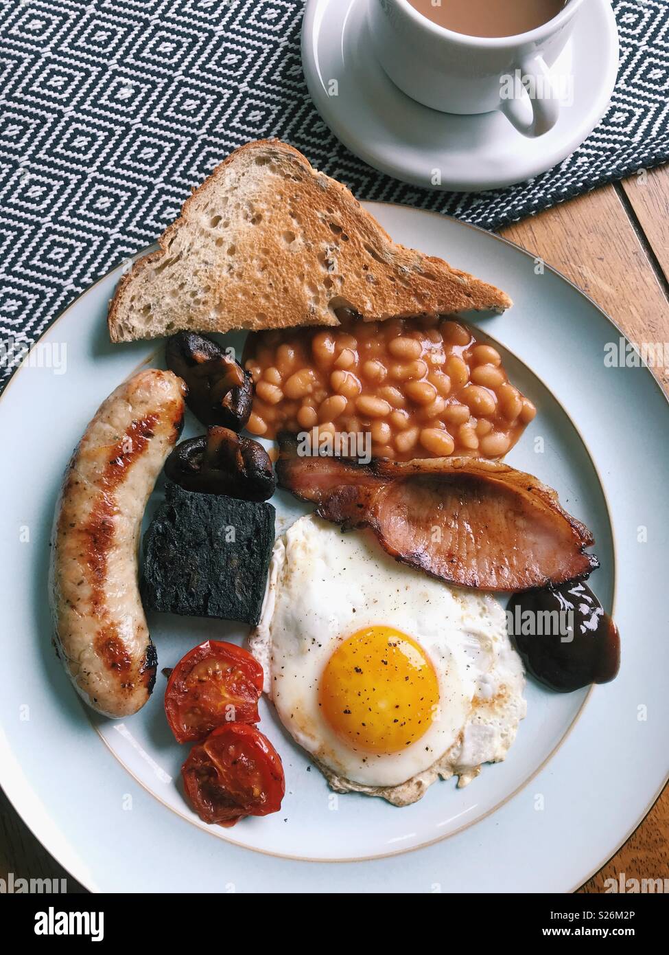 Overhead view of a homemade full English breakfast, with sausage, tomatoes, black pudding, mushrooms, baked beans, toast, brown sauce, a fried egg, and a cup of tea. Stock Photo