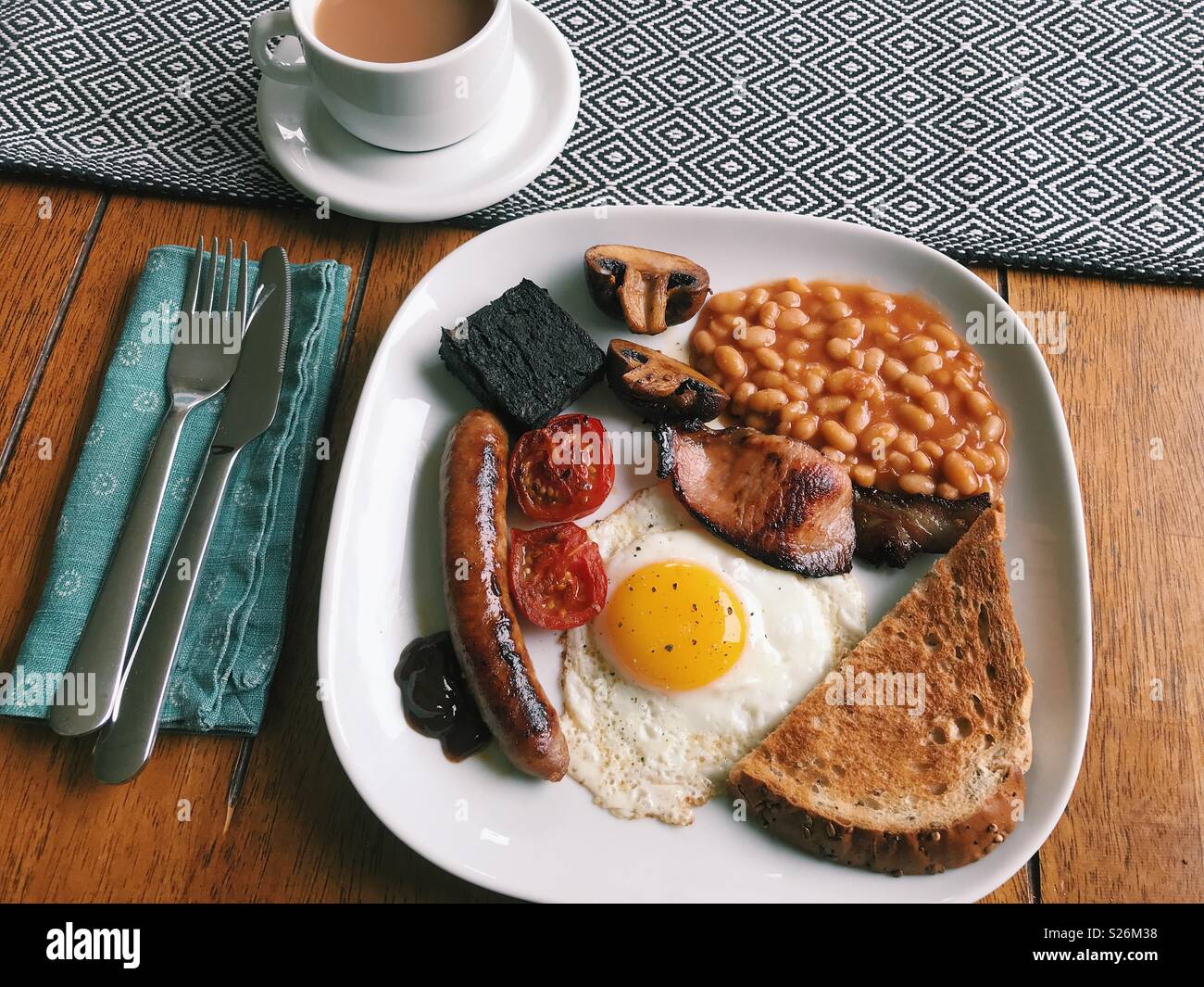 A homemade full English breakfast, with sausage, tomatoes, black pudding, mushrooms, baked beans, toast, brown sauce, a fried egg, and a cup of tea. Stock Photo