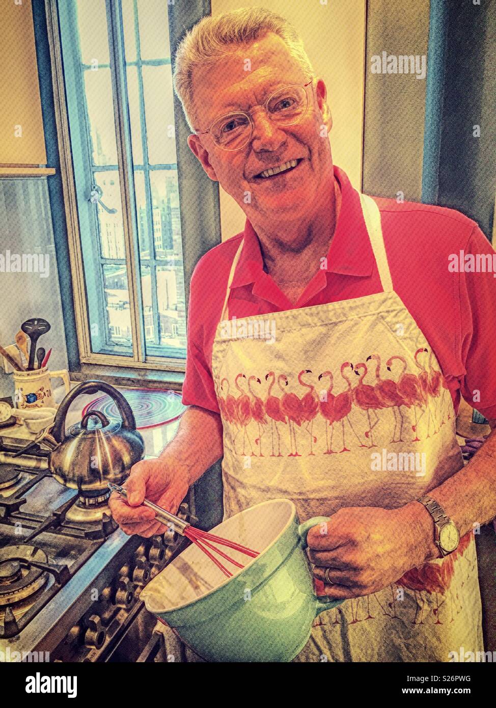 Senior man in apartment kitchen brightly dressed with a flamingo apron, USA - Smartphone Captured Stock Image