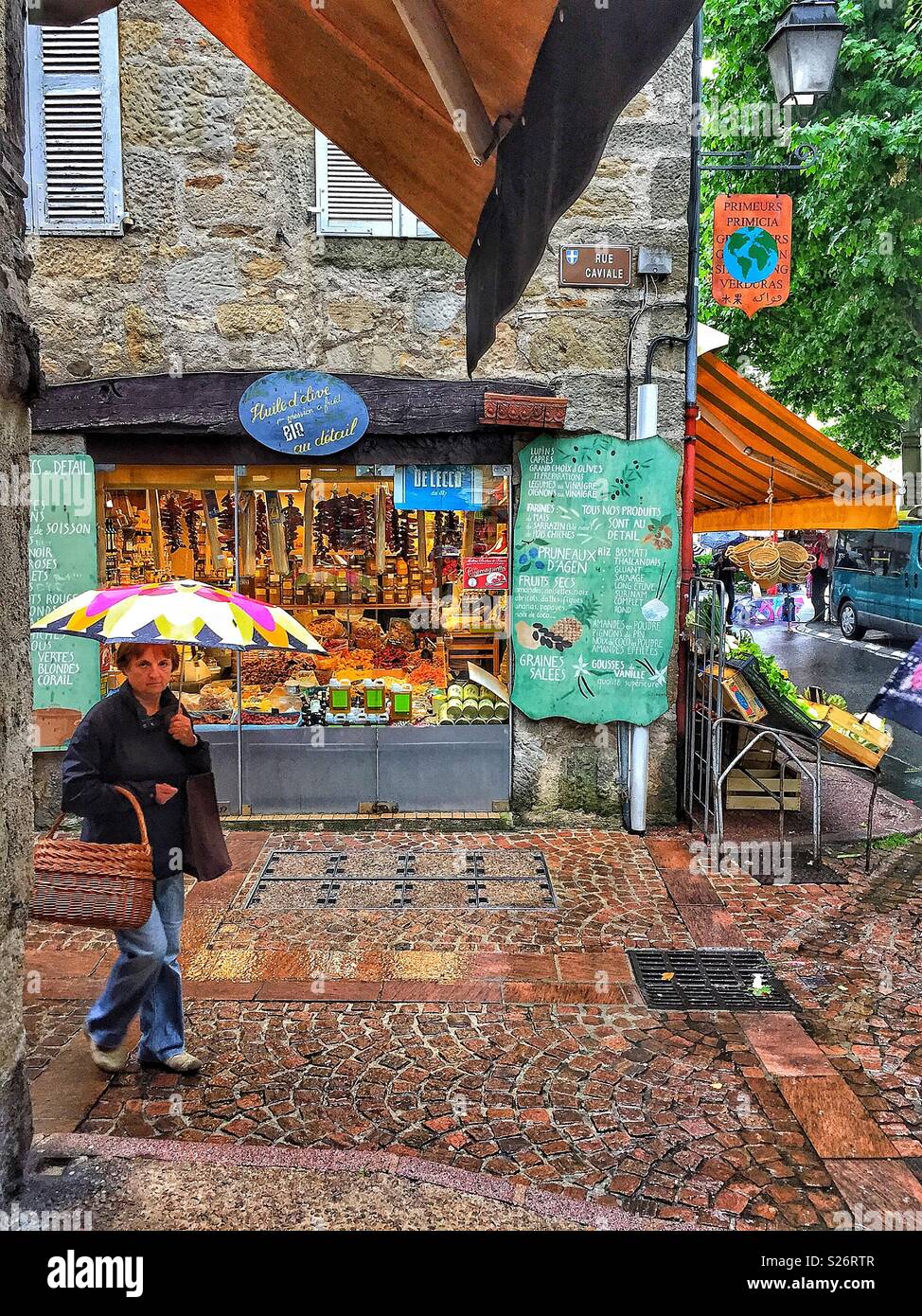 A rainy day in Figeac, France. A small market town in the heart of the Lot Department. A lady is shopping for fruit and vegetables, carrying an umbrella and traditional basket. - Smartphone Captured Stock Image