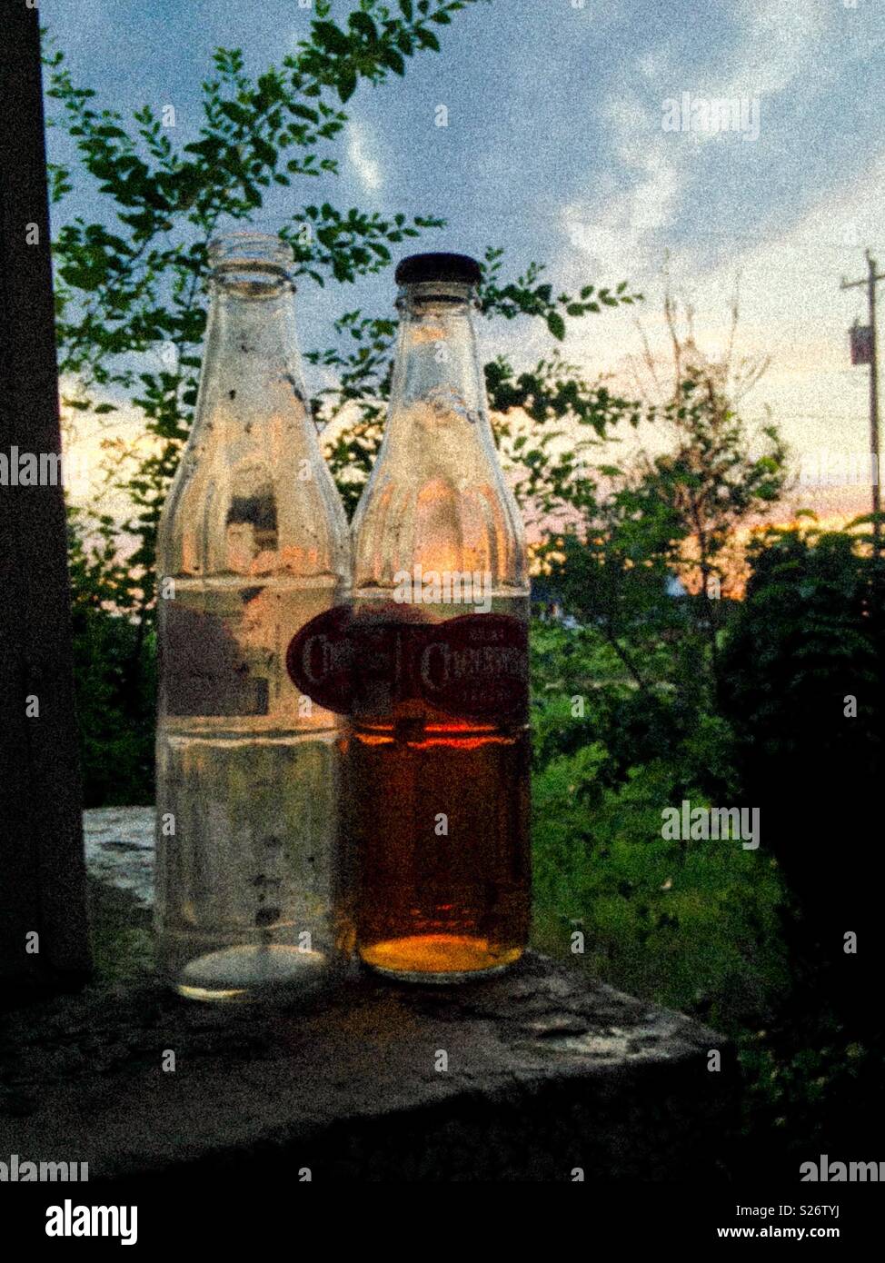 Grainy photo of 2 Cheerwine bottles on the porch in setting sun, one bottle is unopened and evaporated - Smartphone Captured Stock Image