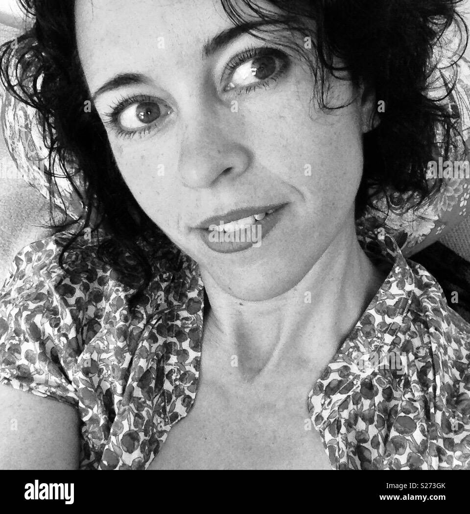 Black and white of women with dark wavy hair in her thirties looking sideways and smiling softly with a short sleeved floral top on - Smartphone Captured Stock Image