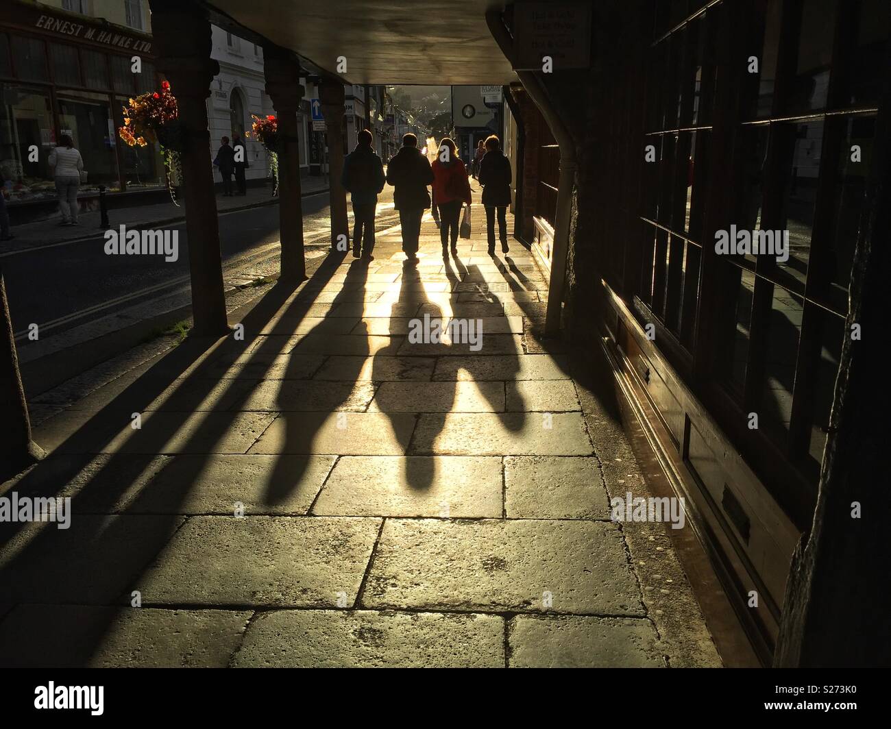 Four pedestrians walk towards the evening sun, causing long shadows to fall on the pavement. Dartmouth, Devon, England, U.K. - Smartphone Captured Stock Image