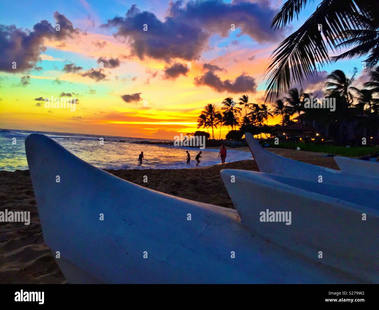 Sunset beach goers frolic in the tropical island waves under the swaying palm trees as outrigger canoes rest until morning - Smartphone Captured Stock Image
