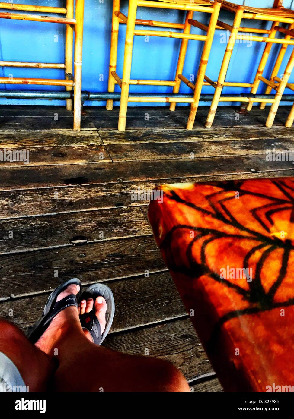 Sandal clad tanned male legs chilling out in a rustic tropical bar with wooden floorboards and bamboo barstools - Smartphone Captured Stock Image
