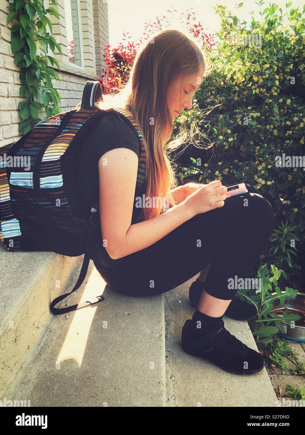 Tween schoolgirl with long blonde hair sitting on step wearing backpack looking at smartphone - Smartphone Captured Stock Image