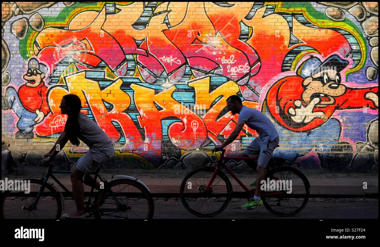 2 cyclists ride past a colourful piece of street art painted on the side of a large wall. It’s summer somewhere in Europe. Photo Credit - © COLIN HOSKINS. - Smartphone Captured Stock Image