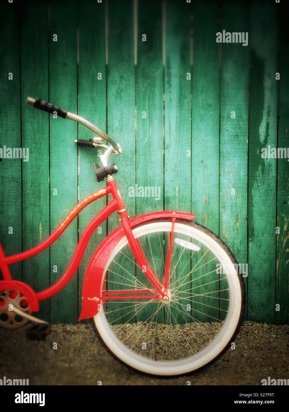 Front half of retro red Flyer bicycle with white tires, parked against a green painted wood fence on a gravel path. - Smartphone Captured Stock Image