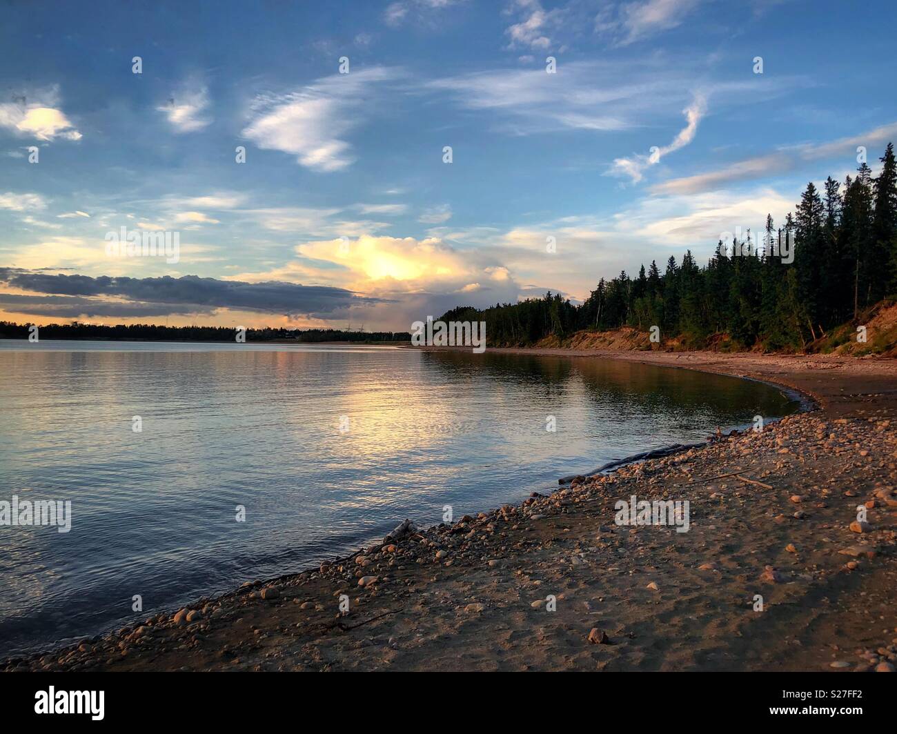 Sunset on Alberta lake with rocky and sandy beach. Spruce treeline in background. Calm blue waters with bright blue sky behind. Few clouds. Stock Photo