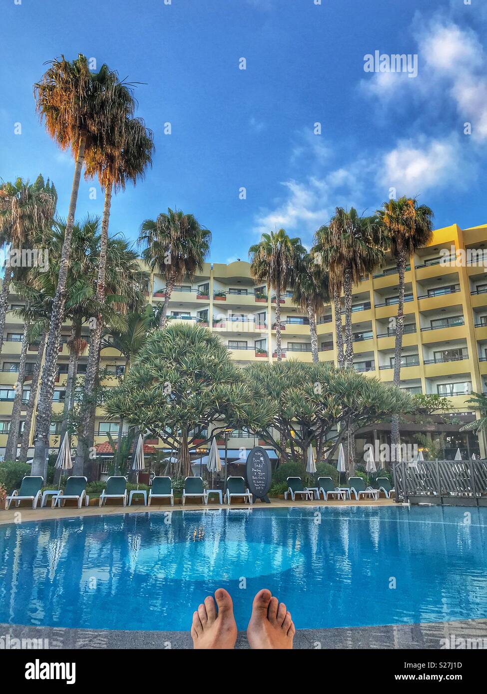 POV lying on a deckchair, looking to bare feet with swimming pool and hotel gardens and building beyond. Funchal, Madeira, Portugal Stock Photo