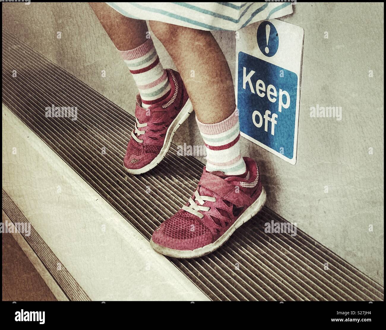 A girl rests her feet on a ledge that is also a radiator. The sign says “Keep Off!” She’s ignoring the request. Children never listen?! Photo Credit - © COLIN HOSKINS. - Smartphone Captured Stock Image