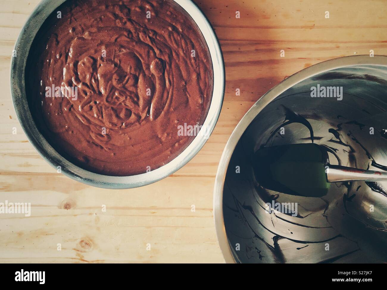 Flat lay view of chocolate cake batter poured into a circular cake pan with empty mixing bowl and spatula to the side on a wooden table - Smartphone Captured Stock Image