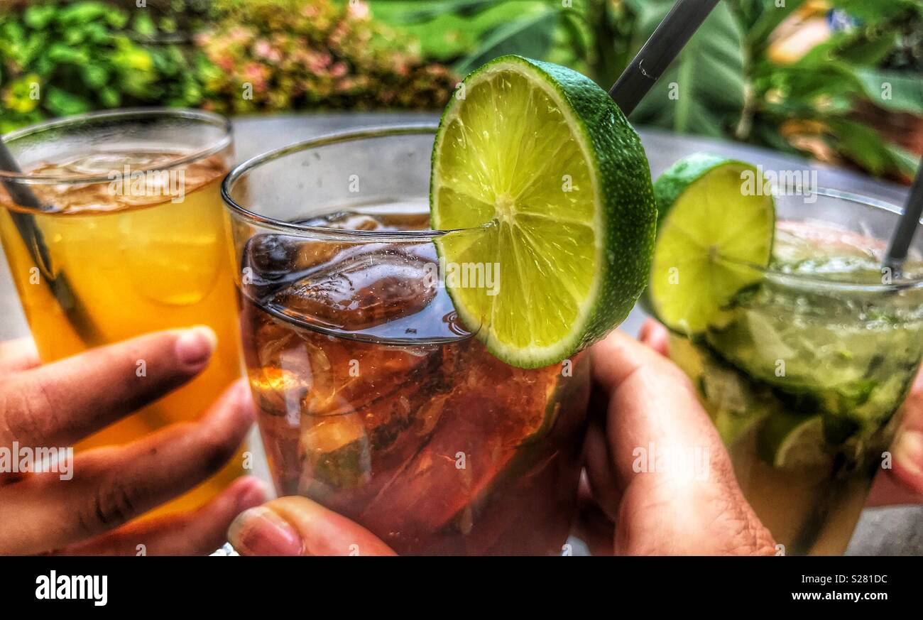 Happy Hour, cocktails, Mojito, rum and coke, poncha, all with ice and a slice of lime, in women’s hands, at a table outdoors in Funchal, Madeira, Portugal Stock Photo
