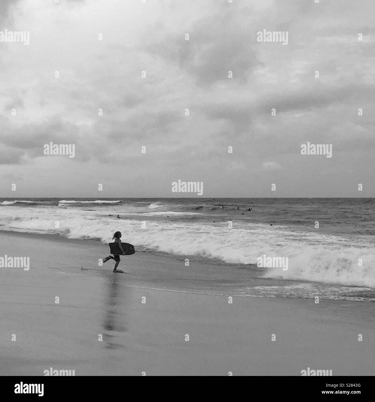 Person carrying a boogie board running towards the ocean. Black and white edit. Square crop. - Smartphone Captured Stock Image