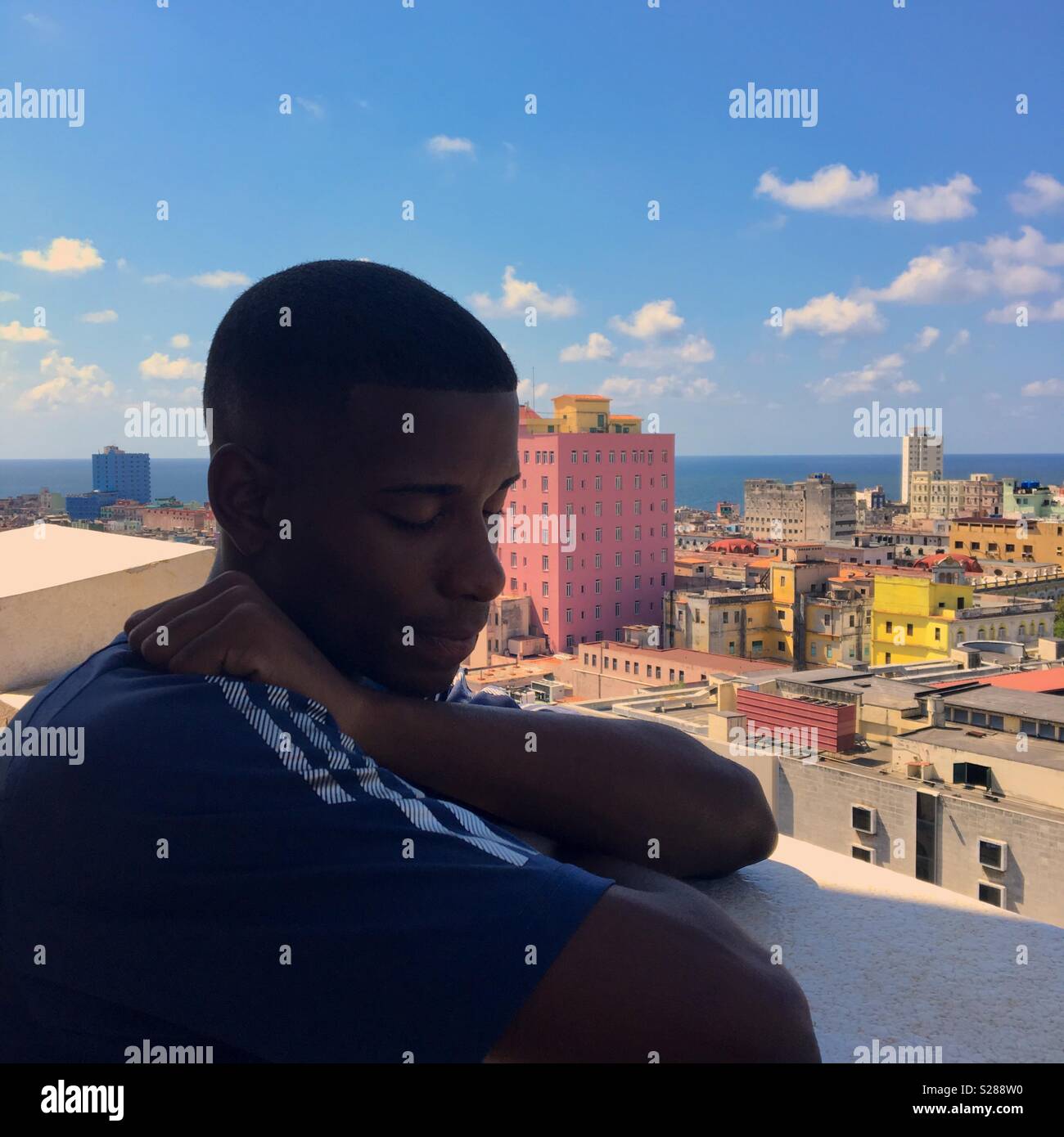 Local Cuban man on a balcony looking sad closing his eyes with Havana buildings and sea in background - Smartphone Captured Stock Image