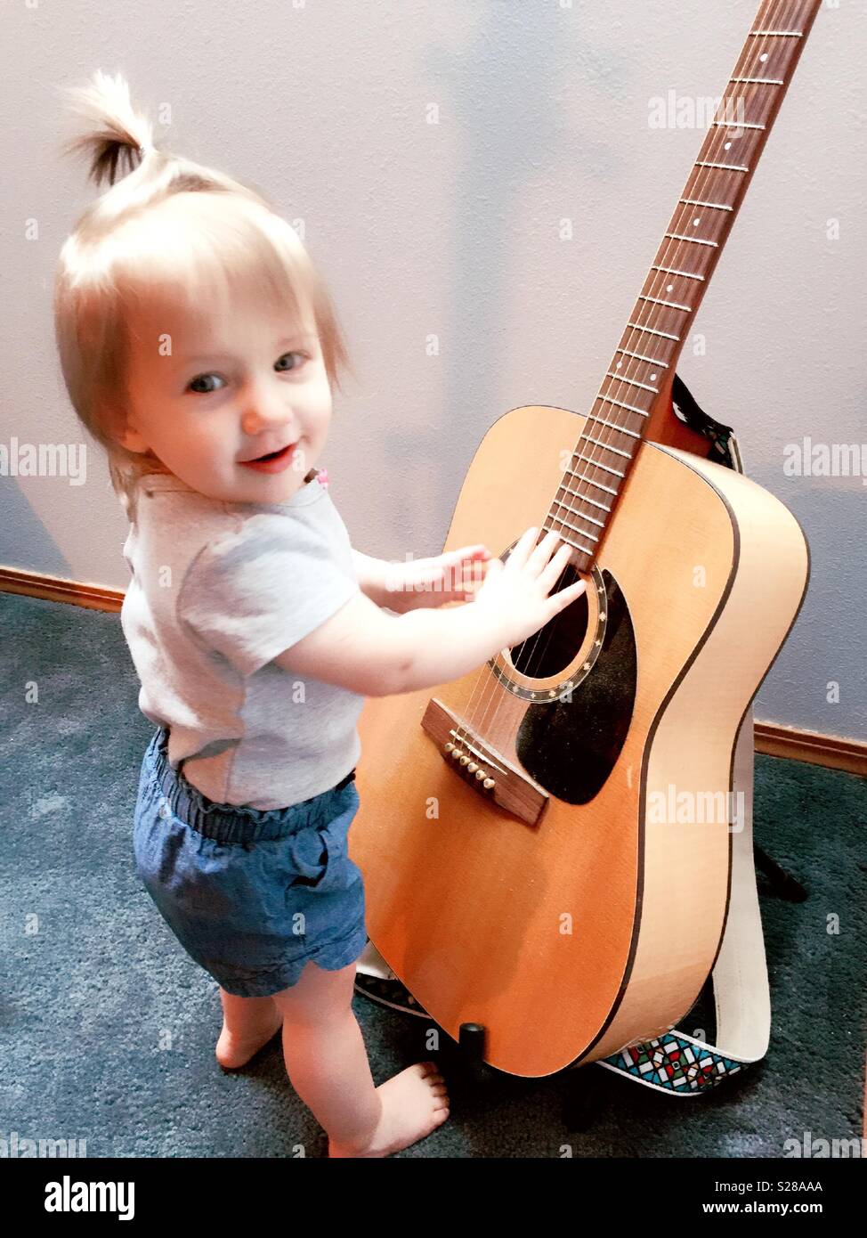 One and a half-year-old toddler reaching out to a acoustic guitar on a stand in her house, USA Stock Photo