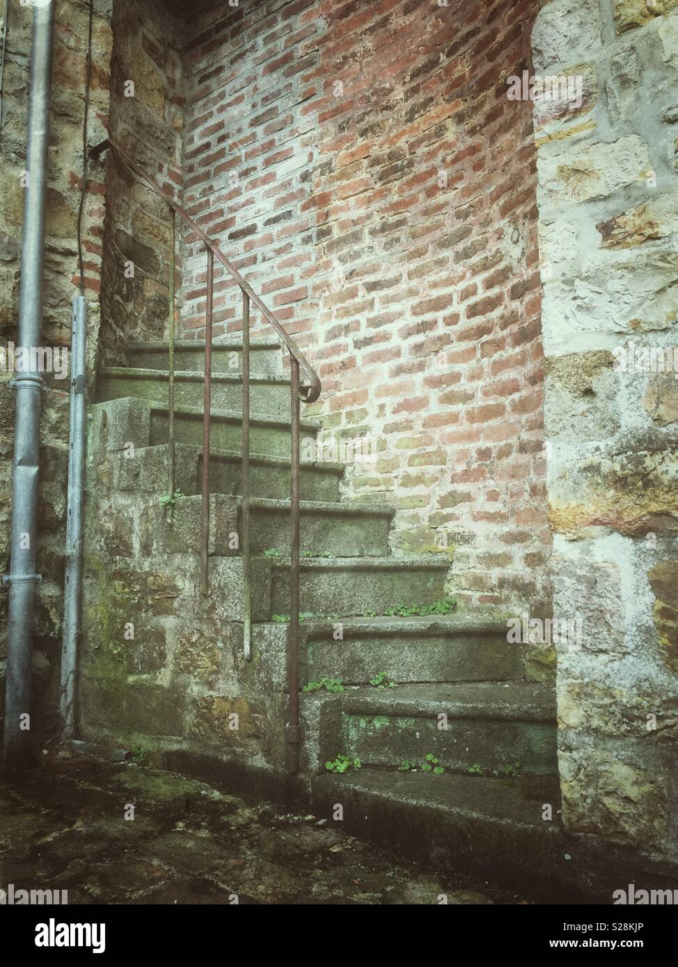Stairs to nowhere. An old, weathered staircase with moss-covered steps and a metal handrail leads up and into a brick wall, going nowhere - Smartphone Captured Stock Image