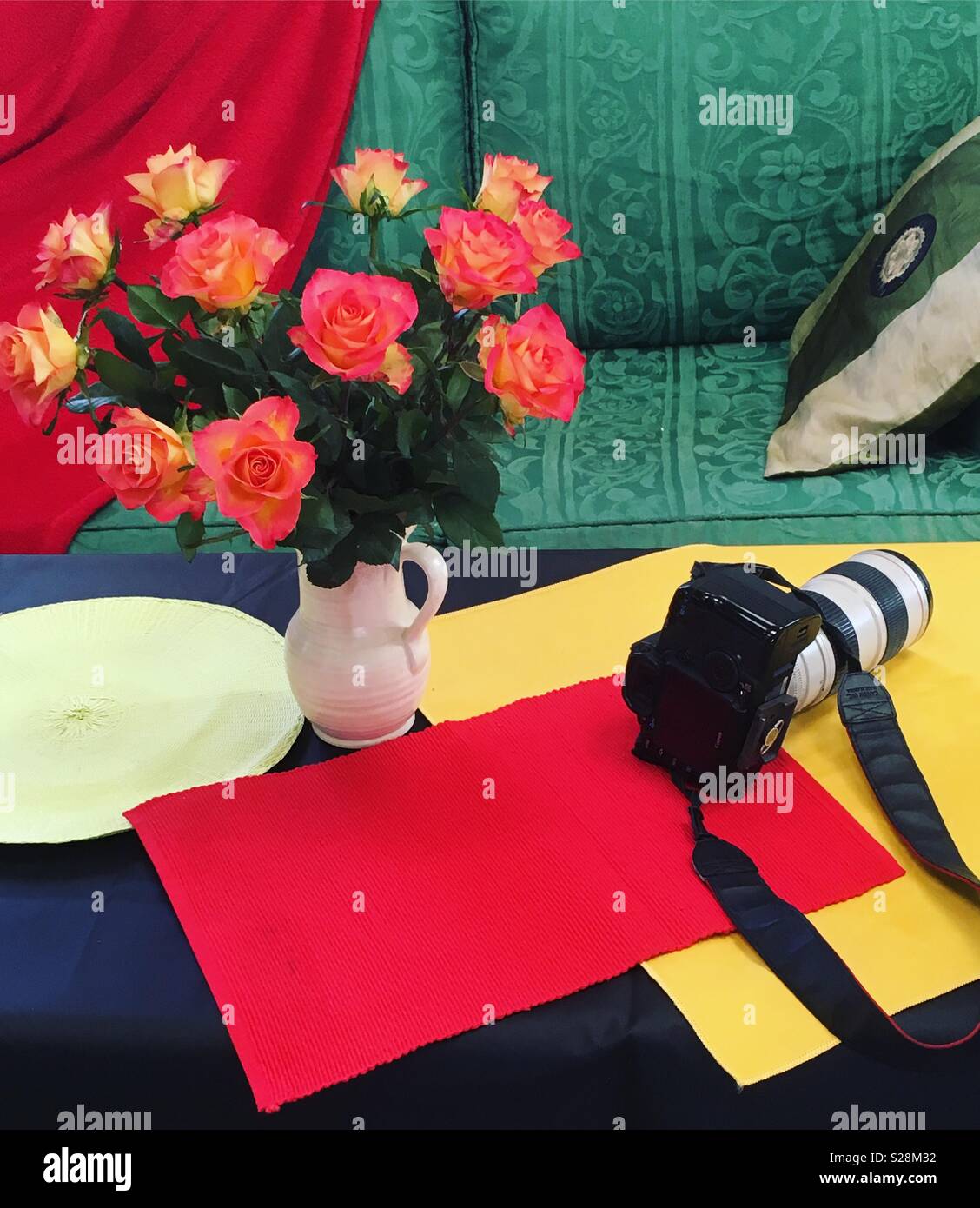 Red and yellow roses in vase on table with colourful placement mats and Canon camera. Stock Photo