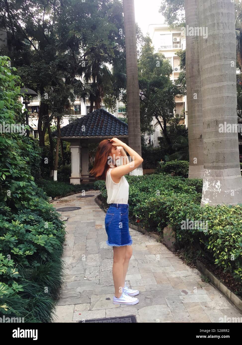 A stylish or fashionable Chinese or Asian girl / lady / woman / female in white blouse, denim skirt and white Converse trainers stroking her brown hair in a garden, photo taken in Guangzhou, China Stock Photo