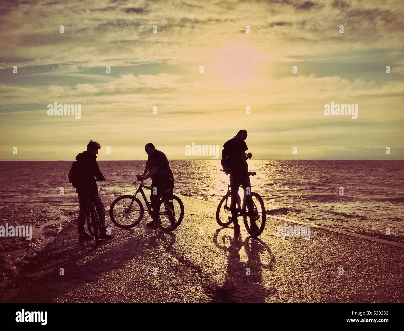 The silhouettes of three male cyclist friends as they relax near the edge of the sea at sunset. A Creative & atmospheric picture with multiple potential uses. Photo Credit - © COLIN HOSKINS. - Smartphone Captured Stock Image