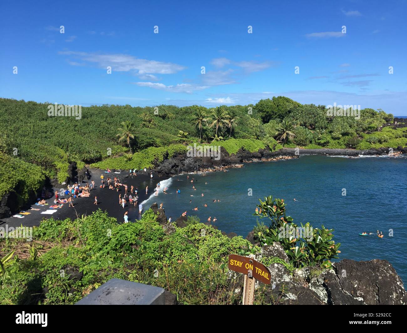 Black sand beach ocean cove with people swimming along jungle hiking path overlook on trail cliff with panoramic rainforest view and Stay on Trail sign along the Road to Hana in Maui Hawaii Stock Photo