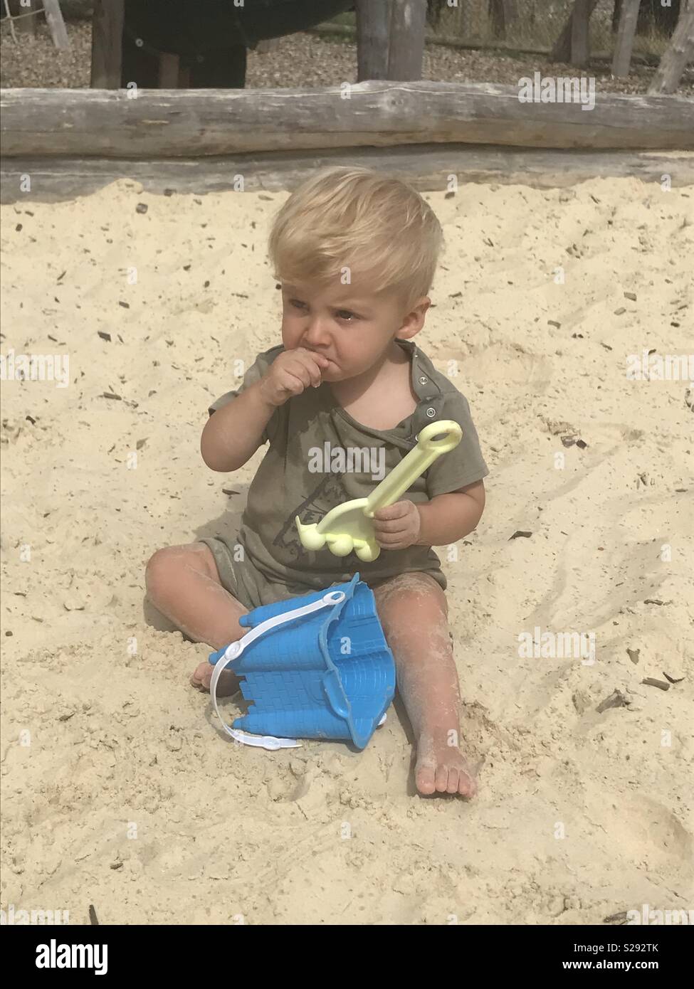 Toddler boy with bucket & spade eating sand in a sandpit Stock Photo