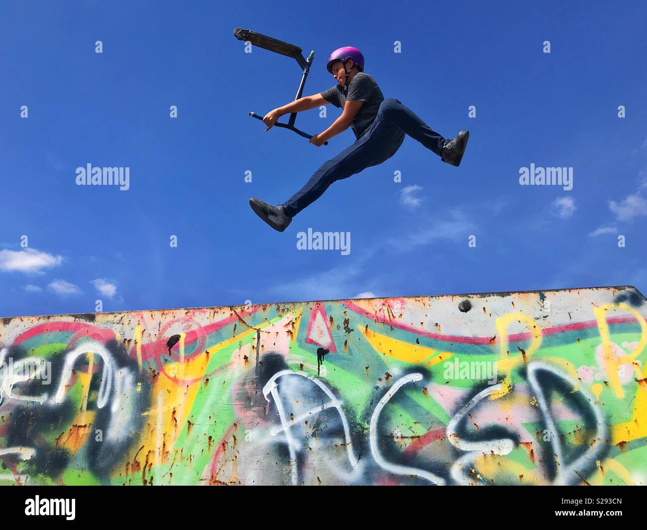 A teenage boy performs a scooter rotation stunt mid air above a graffiti covered ramp. It’s summer and time to get out doors and enjoy the sport of scootering. Photo Credit © COLIN HOSKINS. Stock Photo