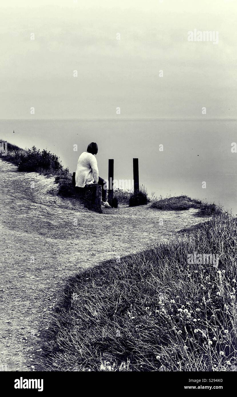 Woman sitting on a cliff top looking out to sea Stock Photo - Alamy