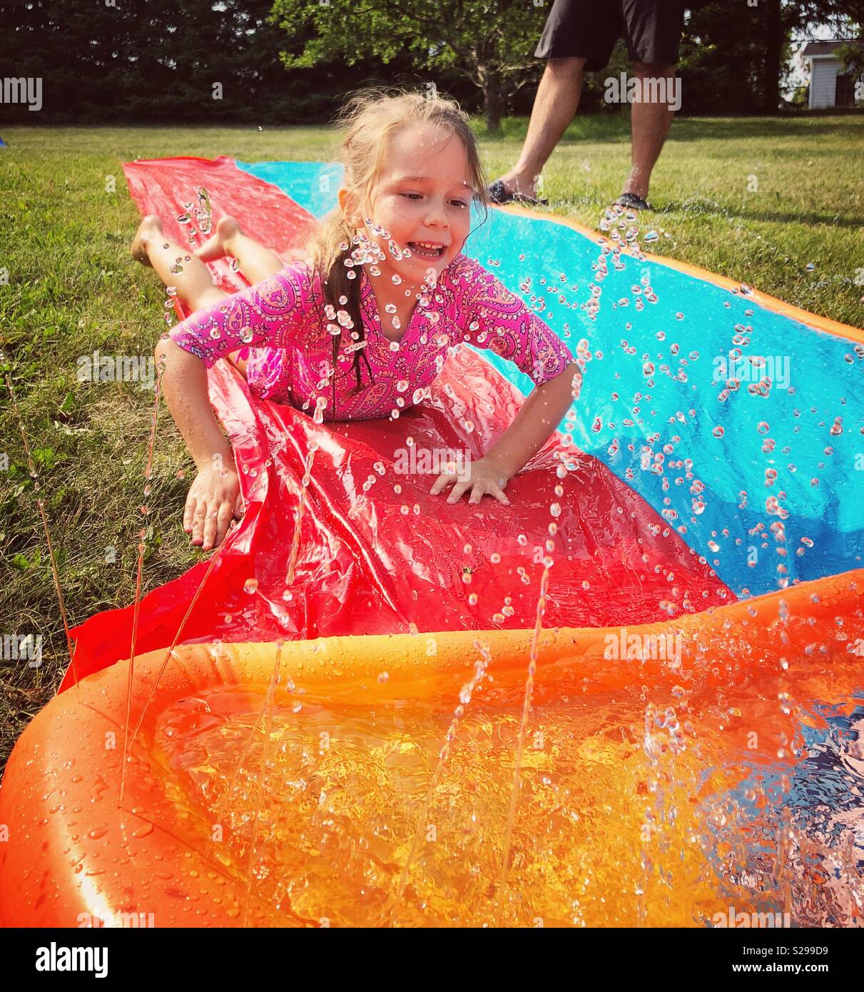 6 year old girl sliding down plastic water sprinkler slide toy outside Stock Photo