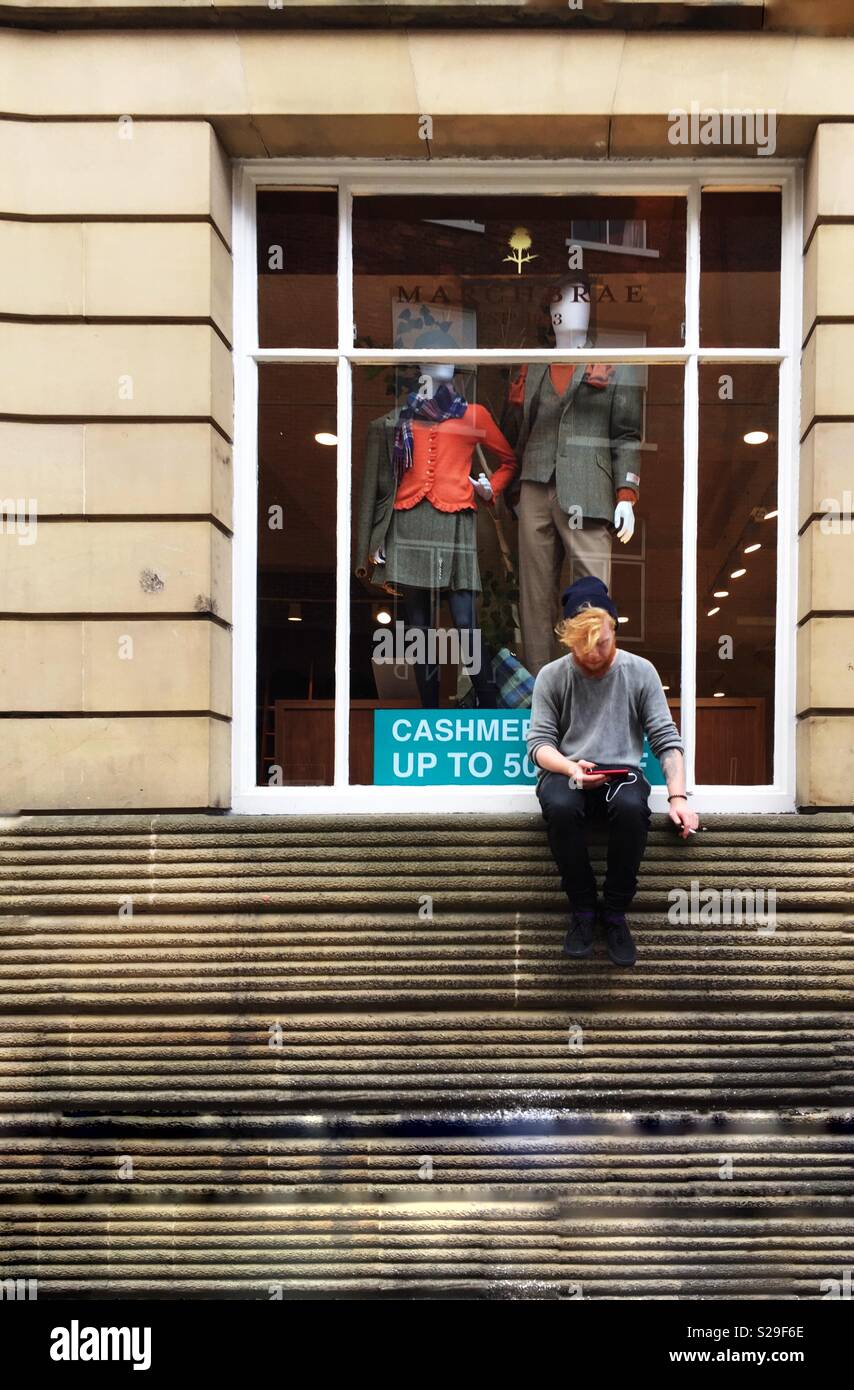 A hipster male perched on a high shop window ledge whilst looking at his mobile phone Stock Photo